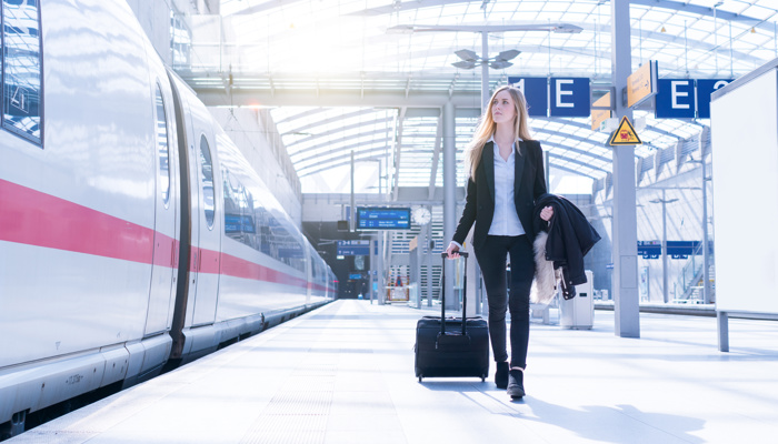 Junge Frau mit Rollkoffer geht am Bahnsteig an einem stehenden Zug entlang.